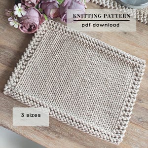 May include: A rectangular beige knitted piece with a thick braided border. The text on the image reads KNITTING PATTERN pdf download and 3 sizes. The item is displayed next to several artificial purple and white flowers.