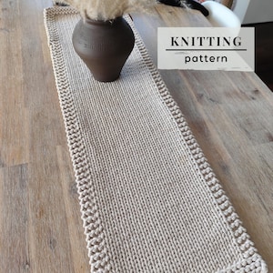 Puede incluir: Un camino de mesa rectangular de punto beige con un borde texturizado y un panel central liso. Un jarrón marrón oscuro con follaje seco se sitúa sobre el camino. La imagen incluye el texto KNITTING pattern en un recuadro blanco.