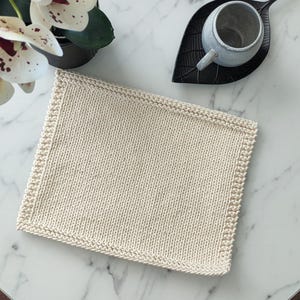 Op de afbeelding: Cr&egrave;mekleurige, vierkante gebreide washand met een getextureerd oppervlak en een decoratieve rand. De washand ligt op een witte marmeren tafel, naast een kopje en een decoratief blad.