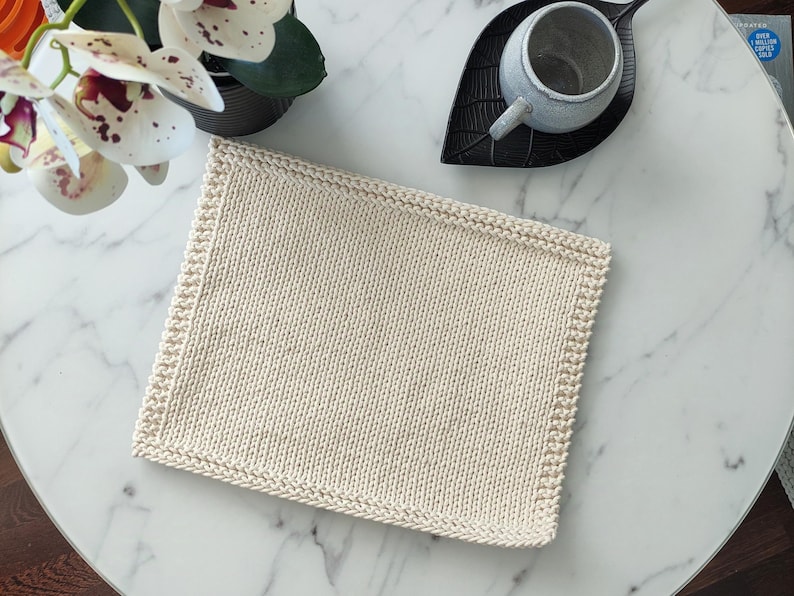Op de afbeelding: Cr&egrave;mekleurige, vierkante gebreide washand met een getextureerd oppervlak en een decoratieve rand. De washand ligt op een witte marmeren tafel, naast een kopje en een decoratief blad.