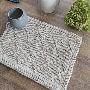 May include: A beige knitted crochet placemat with a diamond pattern. The placemat is made of thick yarn and has a textured surface.