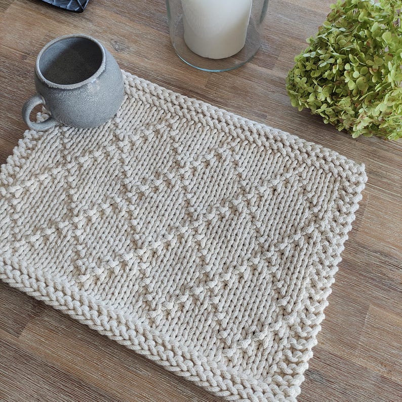 May include: A beige knitted crochet placemat with a diamond pattern. The placemat is made of thick yarn and has a textured surface.