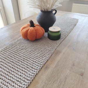 Könnte beinhalten: Ein beigefarbener, gestrickter Tischläufer mit texturiertem Strickmuster. Auf dem Läufer stehen ein flauschiger orangefarbener Kürbis mit braunem Stiel, eine mattschwarze Keramikvase mit hohen Trockengräsern und ein grünes Glasgefäß mit Holzdeckel.