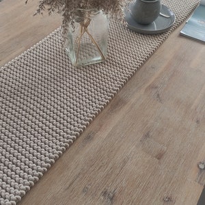 May include: A beige textured table runner with a cup and saucer, and a vase of dried flowers. The runner is made of a woven material and sits on a light-coloured wooden table. A magazine is also on the table.