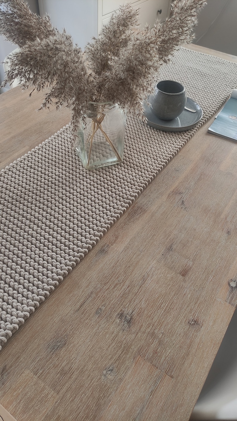 May include: A beige textured table runner with a cup and saucer, and a vase of dried flowers. The runner is made of a woven material and sits on a light-coloured wooden table. A magazine is also on the table.