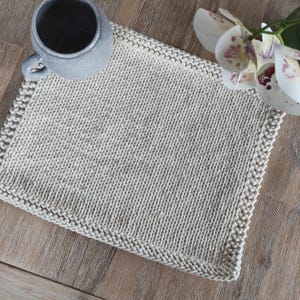 Op de afbeelding: Een beige, handgebreide placemat met een getextureerd oppervlak en een decoratieve rand. Een grijze mok met een handvat staat op de placemat. De placemat ligt op een houten oppervlak. Een witte en paarse orchidee bloem staat op de achtergrond.
