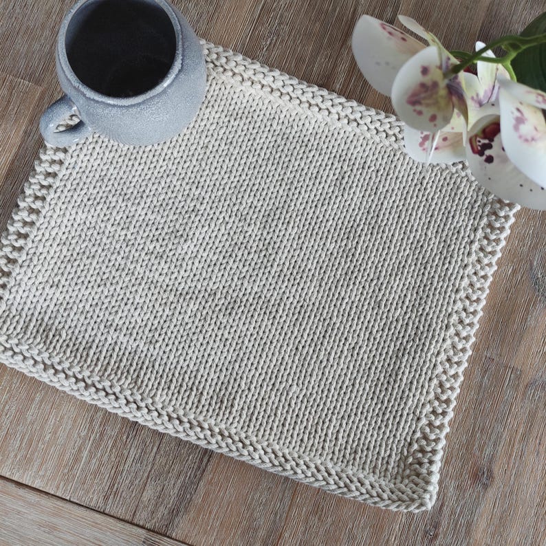 Op de afbeelding: Een beige, handgebreide placemat met een getextureerd oppervlak en een decoratieve rand. Een grijze mok met een handvat staat op de placemat. De placemat ligt op een houten oppervlak. Een witte en paarse orchidee bloem staat op de achtergrond.