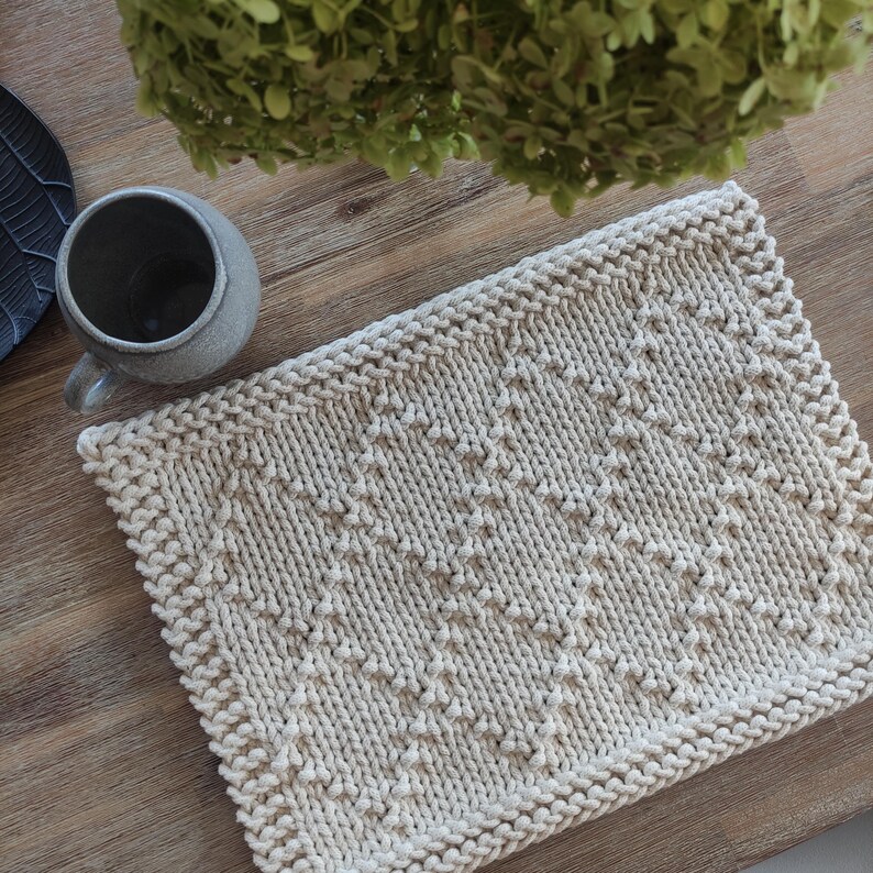 May include: A beige knitted placemat with a diamond pattern. The placemat is made of thick yarn and has a textured surface.