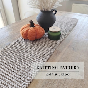 Könnte beinhalten: Ein beigefarbener gestrickter Tischläufer mit strukturiertem Strickmuster. Auf dem Läufer stehen ein flauschiger orangefarbener Kürbis mit braunem Stiel, eine schwarze Keramikvase mit getrockneten Gräsern und ein grünes Glas mit Holzdeckel. Text: KNITTING PATTERN pdf & video.