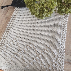 Könnte beinhalten: Ein beigefarbener, gestrickter Tischläufer mit strukturiertem Muster und dekorativem Rand. Der Läufer ist rechteckig und scheint handgefertigt zu sein. Eine Vase mit grünen Blumen steht auf dem Läufer.