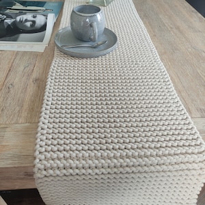 May include: A cream-coloured knitted table runner with a textured pattern. A grey ceramic mug and saucer with a spoon are on the runner. The runner is on a wooden table, creating a neutral, cosy aesthetic.