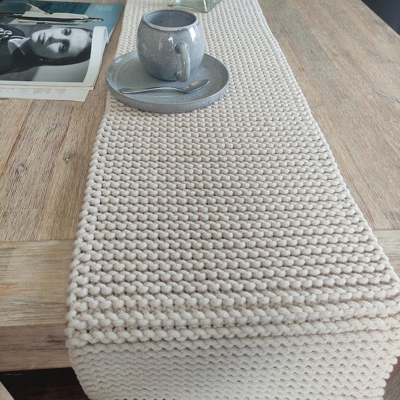 May include: A cream-coloured knitted table runner with a textured pattern. A grey ceramic mug and saucer with a spoon are on the runner. The runner is on a wooden table, creating a neutral, cosy aesthetic.