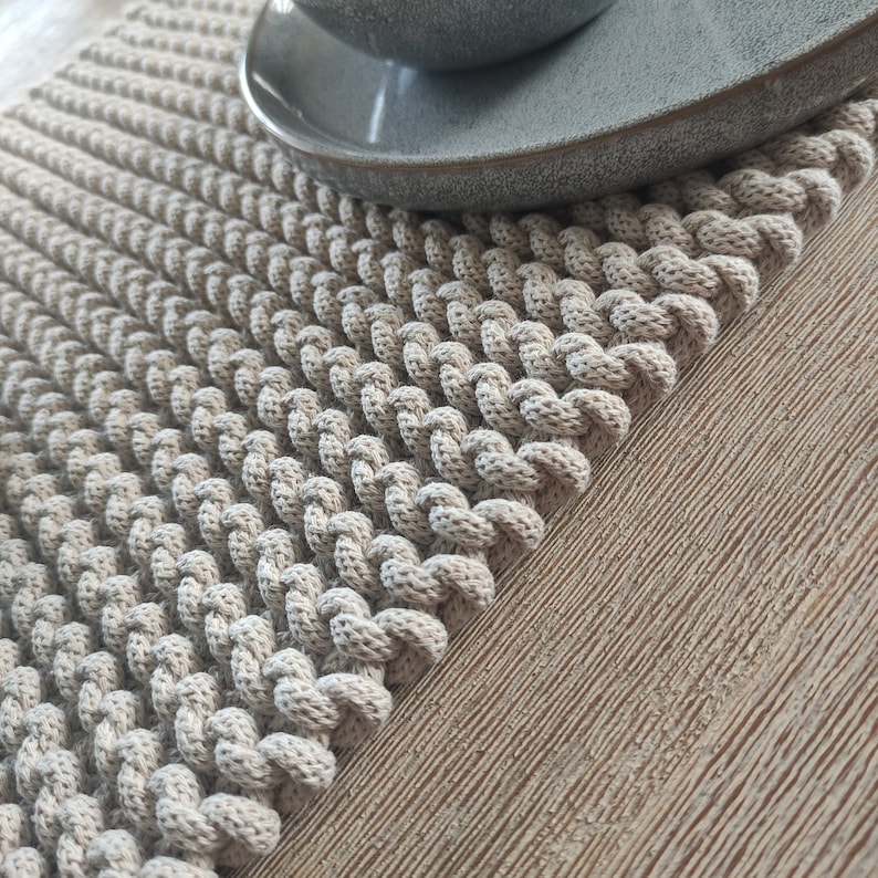 May include: A beige macrame placemat with a textured, braided design. A grey ceramic plate rests on the placemat, which is made of a soft, woven material. The placemat is rectangular and has a decorative edge.