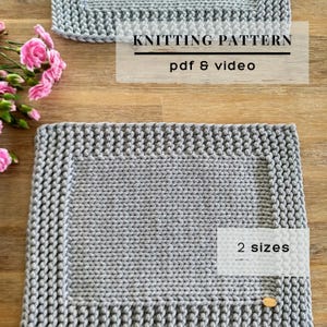 Könnte beinhalten: Ein graues gestricktes Rechteck mit strukturiertem Rand und glatter Mitte. Der Text KNITTING PATTERN pdf & video ist oben sichtbar, und der Text 2 sizes ist unten rechts zu sehen. Rosa Blumen befinden sich auf der linken Seite.