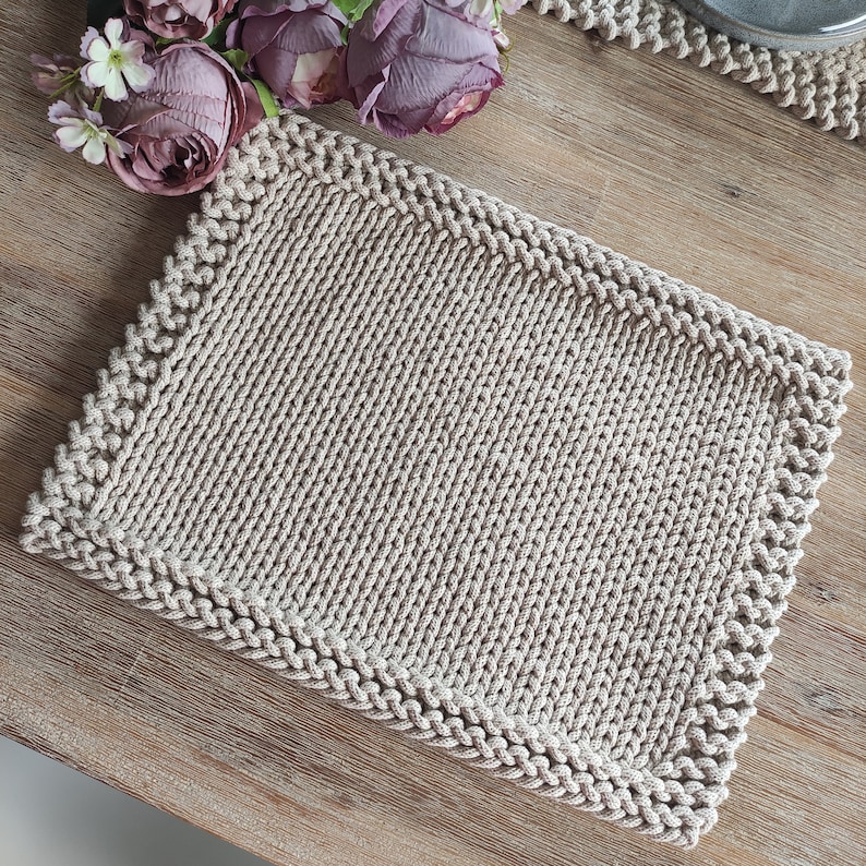 Op de afbeelding: Een rechthoekige, beige gebreide placemat met een getextureerd oppervlak en een decoratieve rand. De placemat wordt weergegeven op een houten tafel, met een boeket paarse bloemen op de achtergrond. De placemat meet ongeveer 30 cm bij 40 cm.
