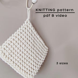 Op de afbeelding: Een vierkante, gebroken witte gebreide pannenlap met een dik, gestructureerd steekpatroon en een ophanglus. De tekst KNITTING pattern pdf & video en 3 sizes staat in zwarte letters naast het item weergegeven.