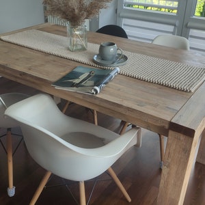 May include: A wooden dining table with a beige runner, a magazine, and a grey teacup and saucer. Two white chairs with wooden legs and grey foot protectors are in front of the table. A vase of dried plants sits on the table.