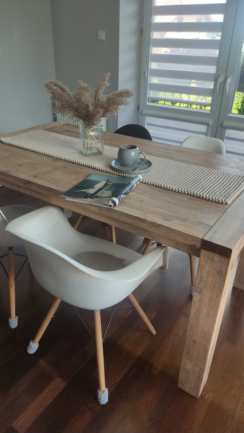 May include: A wooden dining table with a beige runner, a magazine, and a grey teacup and saucer. Two white chairs with wooden legs and grey foot protectors are in front of the table. A vase of dried plants sits on the table.