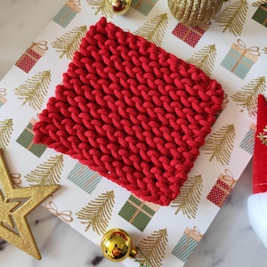 Könnte beinhalten: Ein quadratischer, leuchtend roter Strickuntersetzer mit einer dicken, groben Garnstruktur und einem gleichmäßigen Zopfmuster. Der Untersetzer ist aus einer durchgehenden Kordel gefertigt, was eine dichte und einheitliche Oberfläche mit sichtbaren Schlaufen erzeugt.
