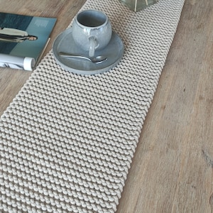 May include: A cream-coloured, hand-knitted table runner with a textured, looped design. A grey ceramic cup and saucer with a spoon sit on the runner. A clear glass vase with dried plants is also on the table. The table has a light wood grain.