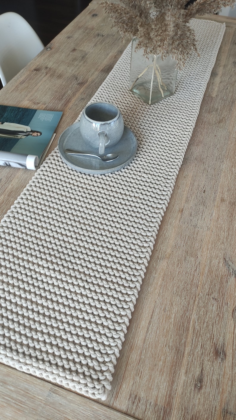 May include: A cream-coloured, hand-knitted table runner with a textured, looped design. A grey ceramic cup and saucer with a spoon sit on the runner. A clear glass vase with dried plants is also on the table. The table has a light wood grain.