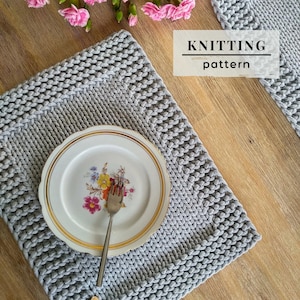 May include: A light gray rectangular knitted placemat with a textured border, featuring a white ceramic plate with a gold rim and a colorful floral pattern in the center. A silver fork rests on the plate. The text KNITTING pattern is visible.