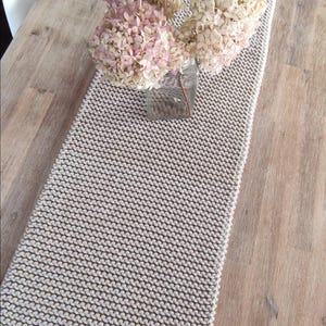 May include: A beige knitted table runner with a textured pattern, placed on a wooden table. A glass jar with dried pink and cream-coloured flowers sits on the runner, adding a touch of natural decor.