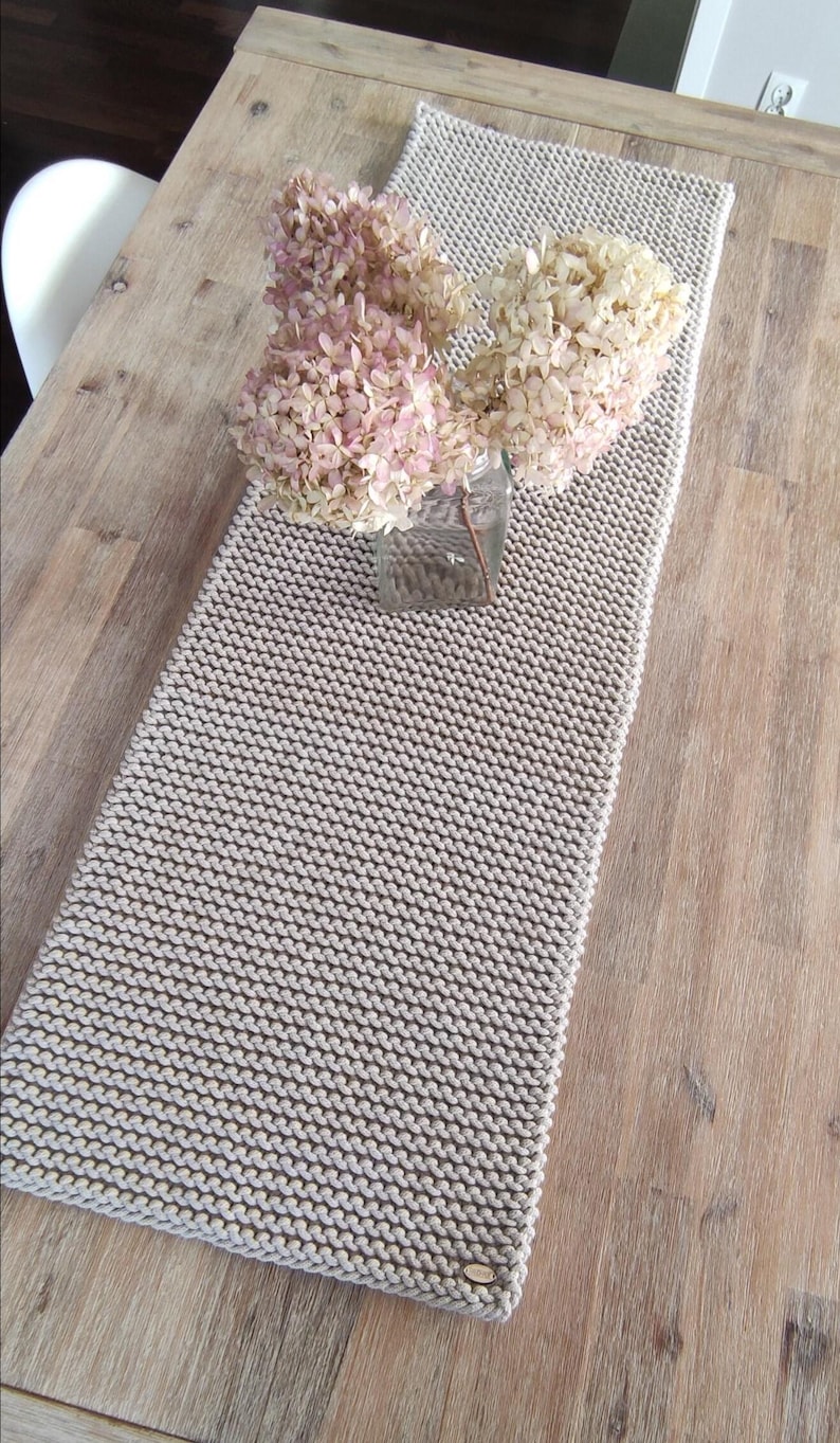 May include: A beige knitted table runner with a textured pattern, placed on a wooden table. A glass jar with dried pink and cream-coloured flowers sits on the runner, adding a touch of natural decor.