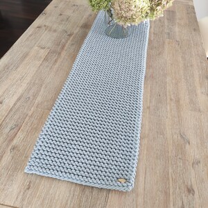 May include: A pale blue knitted table runner with a textured weave. The rectangular runner is laid on a wooden table and features a small wooden tag at one end. A glass vase with flowers sits on the table.