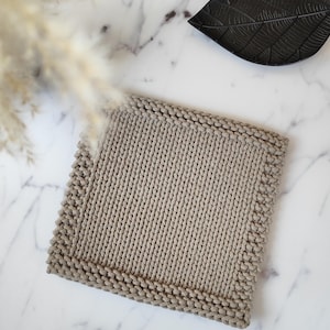 May include: A square, beige knitted washcloth with a textured border. The washcloth is made of a thick yarn and is displayed on a white marble surface. A black leaf-shaped dish is in the background.
