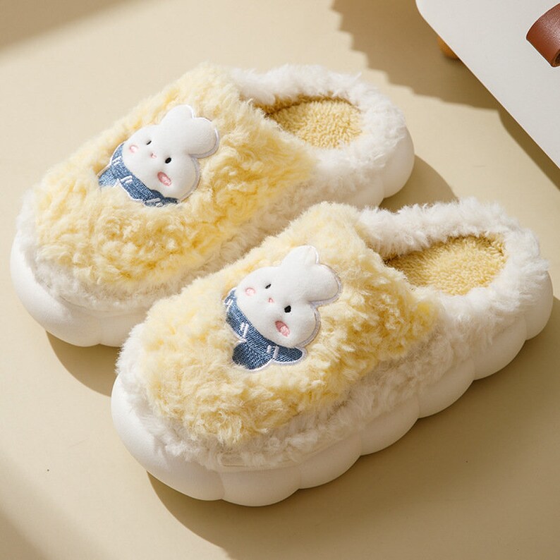 May include: A pair of fluffy yellow and white bunny slippers. Each slipper has a white bunny face with a blue scarf embroidered on the top. The slippers have a soft, plush texture and a white, rounded sole.