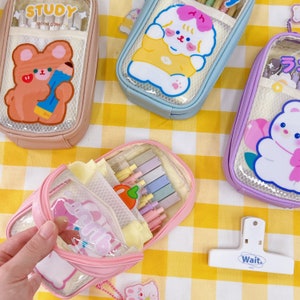 May include: Four clear plastic pencil cases with different cartoon characters on the front. The cases are pink, blue, purple, and pink. The pink case has a bear, the blue case has a dog, the purple case has a bear, and the pink case has a bear. The cases are on a yellow and white checkered background.