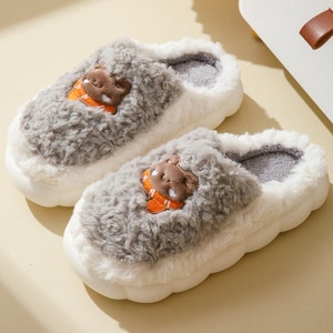 May include: A pair of plush, white and gray slippers with a cartoon bear applique. The slippers have a thick, white sole and a soft, fuzzy texture. The bear applique is brown with an orange scarf.
