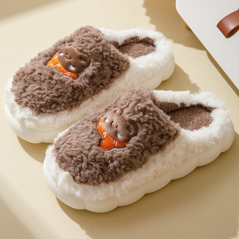May include: A pair of plush, brown and white slippers. Each slipper features a brown bear face with an orange scarf embroidered on the top. The slippers have a fluffy, textured appearance and a thick, white sole.