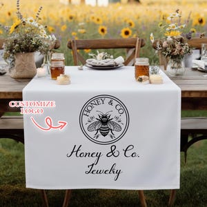 May include: White table runner with a black and white bee logo that says "Honey & Co. Jewelry". The runner is on a wooden table set for a meal, with flowers and jars of honey. The background is a field of sunflowers.