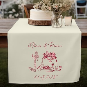 May include: Cream-colored table runner with a burgundy illustration of a floral arrangement, food, and wine bottles. The names "Olivia & Kevin" and the date "11.09.2025" are also printed on the runner. A wedding cake and flowers are on the table.