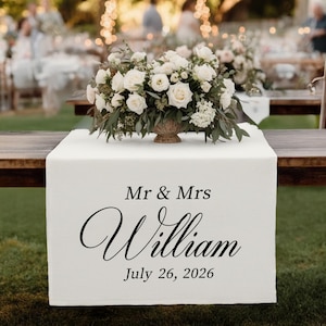 May include: A white table runner with the text "Mr & Mrs William July 26, 2026" in black script. A floral arrangement of white roses and greenery sits atop the runner. The setting appears to be an outdoor wedding reception.