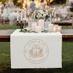 May include: A white table runner with a gold logo that reads "LICERIA & CO. HANDMADE CANDLES" is displayed on a wooden table. The table is decorated with white candles in glass holders and a bronze candelabra. The background is blurred.