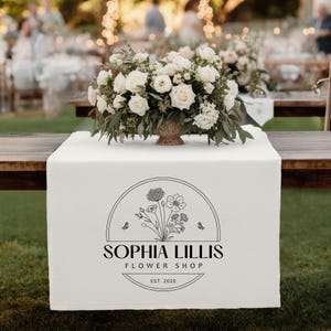 May include: A white table runner featuring the text "SOPHIA LILLIS FLOWER SHOP EST. 2020" and a floral design. A floral arrangement of white roses and greenery sits on the runner. The scene is set outdoors with a blurred background.