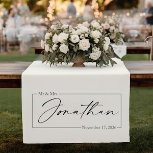May include: A white table runner with the text "Mr & Mrs. Jonathan November 17, 2026" in black script. A floral arrangement of white roses and greenery sits atop the runner. The scene is set outdoors at a wedding reception.