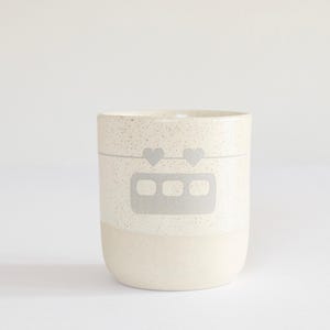Könnte beinhalten: Eine weiße Keramiktasse mit einem gesprenkelten Überzug. Die Tasse hat ein graues Design eines Zuges mit Herzen, die daran hängen.