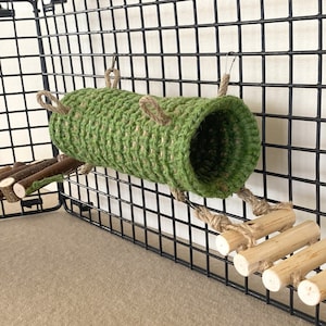 May include: A green crocheted cylindrical tunnel for small animals, suspended by rope loops from a black wire grid panel. A wooden ladder made of light-colored cylindrical rungs connected by rope is attached to the tunnel and the wire grid.