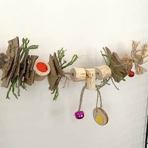 Könnte beinhalten: Ein Vogelspielzeug aus natürlichen Materialien. Es besteht aus Pappe, Holzperlen und Seil, mit Akzenten aus grünem Garn und kleinen Glöckchen. Das Spielzeug ist zum Aufhängen konzipiert und bietet eine Bereicherung für Ziervögel.