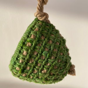 May include: A triangular-shaped ornament crafted from a combination of green and tan natural fiber twine. The piece features a coarse, woven texture and is suspended by a thick, knotted tan rope loop at the top, with a small knot at the bottom corner.