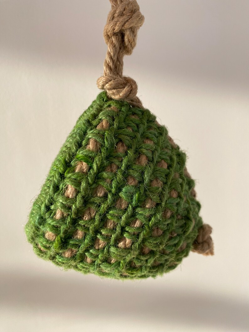 May include: A triangular-shaped ornament crafted from a combination of green and tan natural fiber twine. The piece features a coarse, woven texture and is suspended by a thick, knotted tan rope loop at the top, with a small knot at the bottom corner.