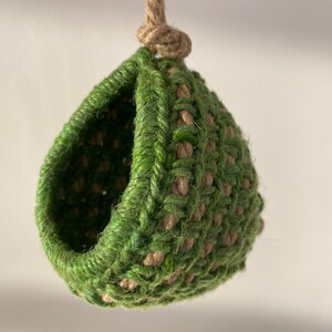 May include: A hanging pod crafted from interwoven green and natural tan fibers. The structure features a rounded, hollow opening and is suspended by a thick, knotted jute rope at the top, creating a textured, spherical shape with a woven pattern.