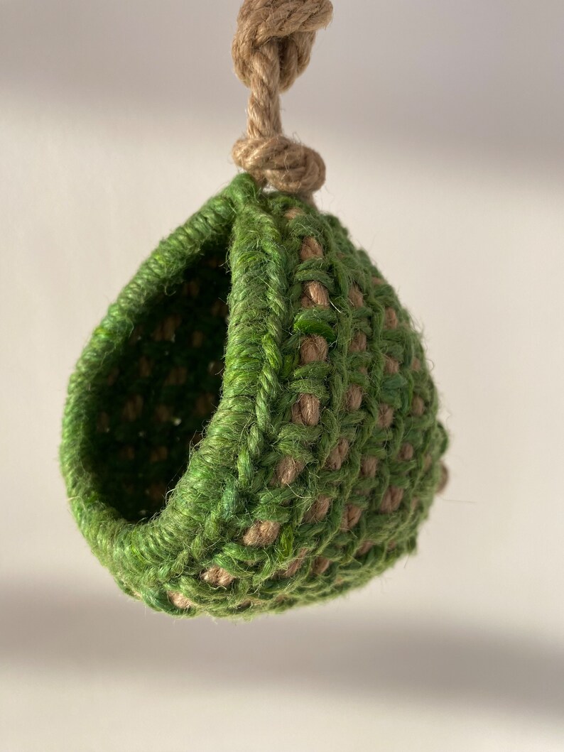 May include: A hanging pod crafted from interwoven green and natural tan fibers. The structure features a rounded, hollow opening and is suspended by a thick, knotted jute rope at the top, creating a textured, spherical shape with a woven pattern.