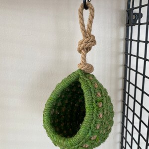 May include: A hanging, teardrop-shaped pod crafted from woven green and natural tan jute fibers. The structure features a wide, circular opening and is suspended by a thick, knotted jute rope attached to a metal carabiner clip at the top.