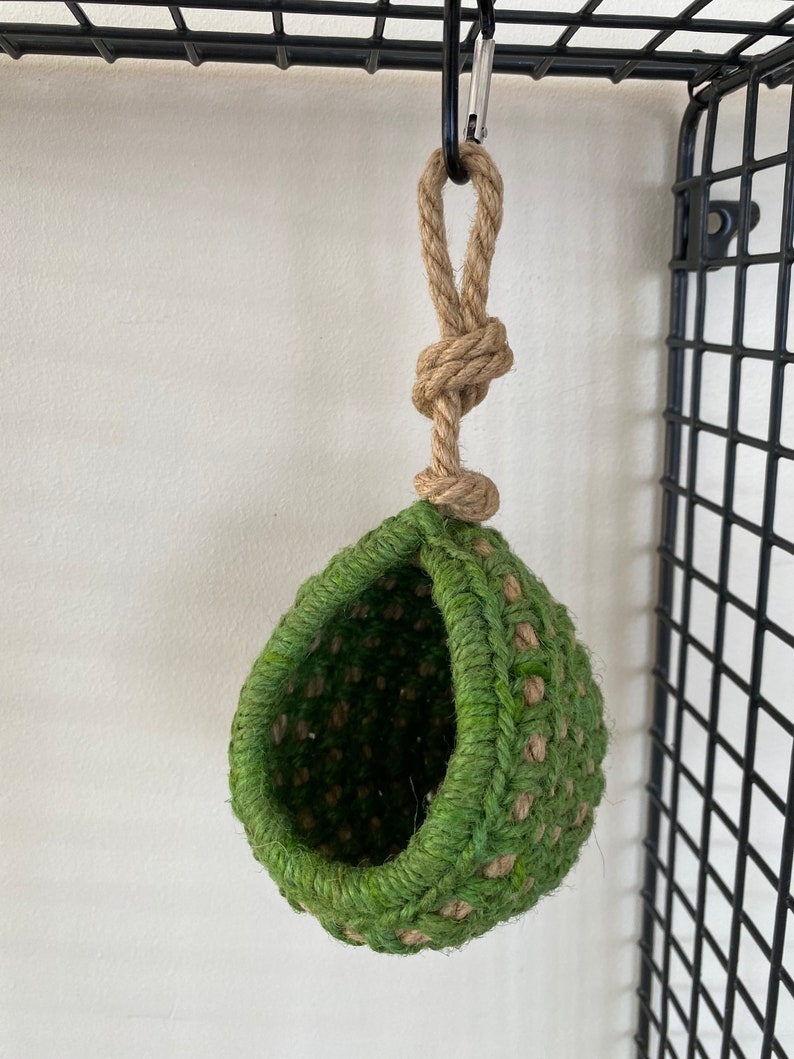 May include: A hanging, teardrop-shaped pod crafted from woven green and natural tan jute fibers. The structure features a wide, circular opening and is suspended by a thick, knotted jute rope attached to a metal carabiner clip at the top.
