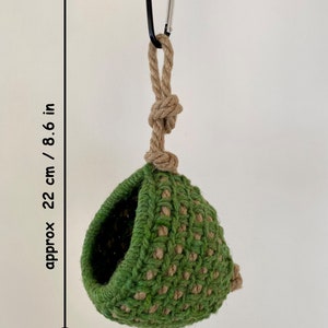 May include: A green and tan crocheted hanging pod with a circular opening, suspended by a thick tan rope and a black metal carabiner. The item measures approximately 8.6 inches in height, as indicated by the text and arrow on the left side.